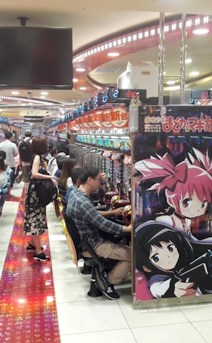 A bustling arcade filled with people playing various gaming machines. Brightly colored displays and animated characters are prominent on posters and screens. The atmosphere is lively with patrons seated in rows, focusing on the games. Overhead signage and electronic scoreboards are visible, contributing to the busy and vibrant environment.